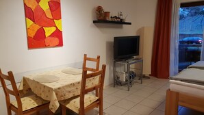 Une pièce comprenant une table à manger, des chaises, un téléviseur posé sur un meuble, une étagère avec divers objets et une fenêtre donnant sur l’extérieur.
