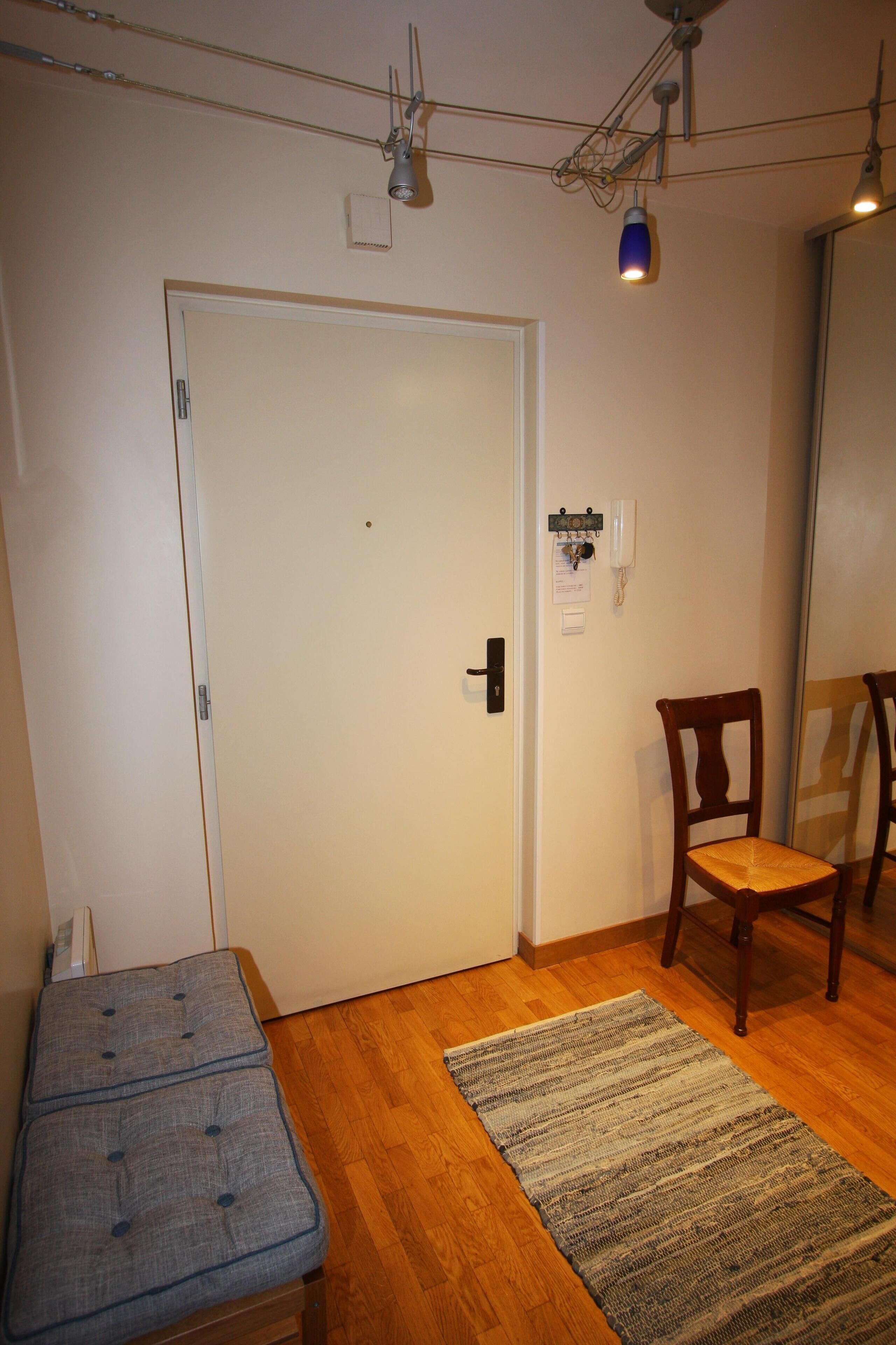 Una habitación con suelo de madera, un banco, una silla, una puerta y una lámpara fijada a la pared.