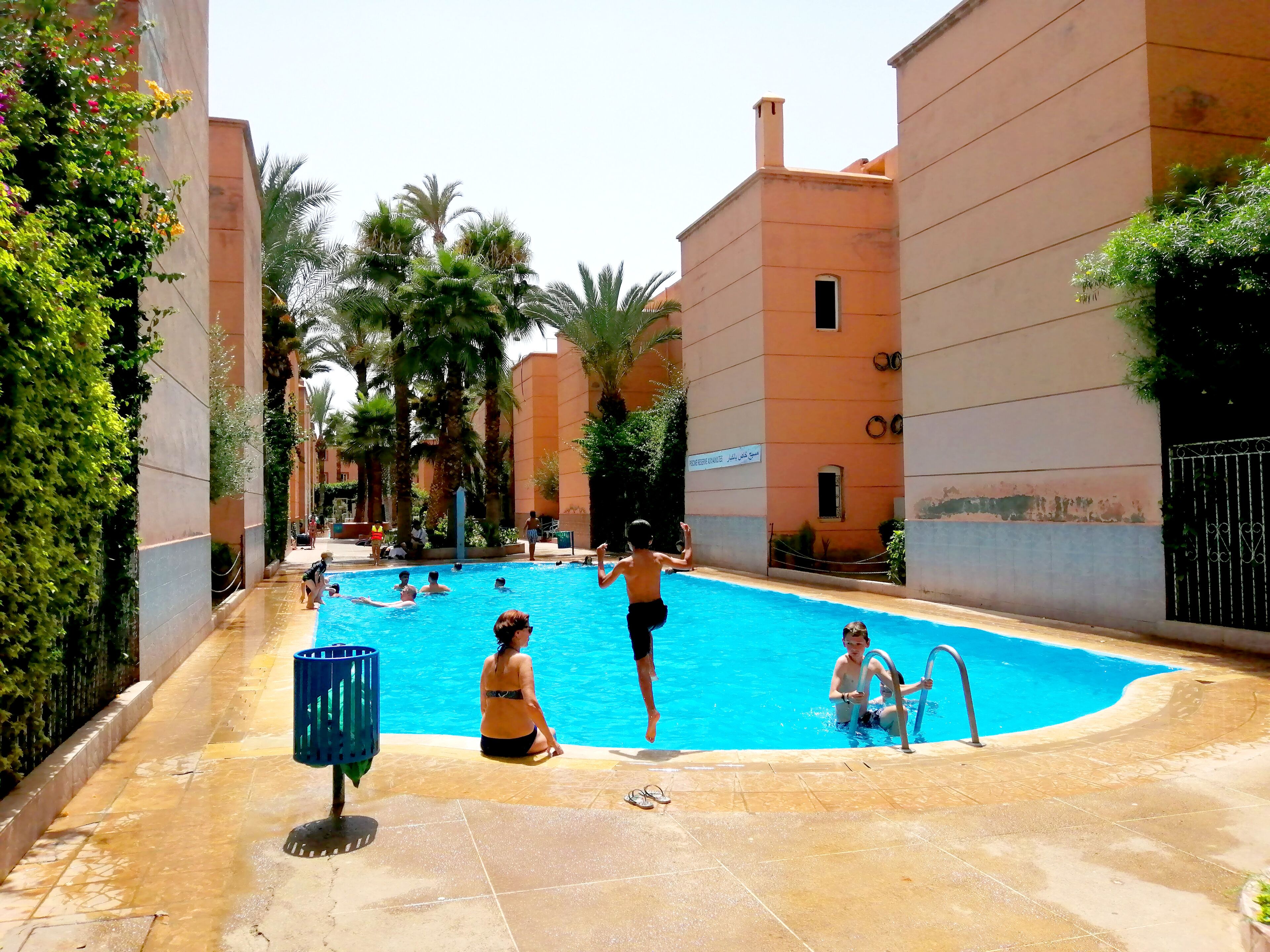 Un espace piscine doté d’un sol carrelé, de palmiers et d’un bâtiment sur lequel est apposée une enseigne.