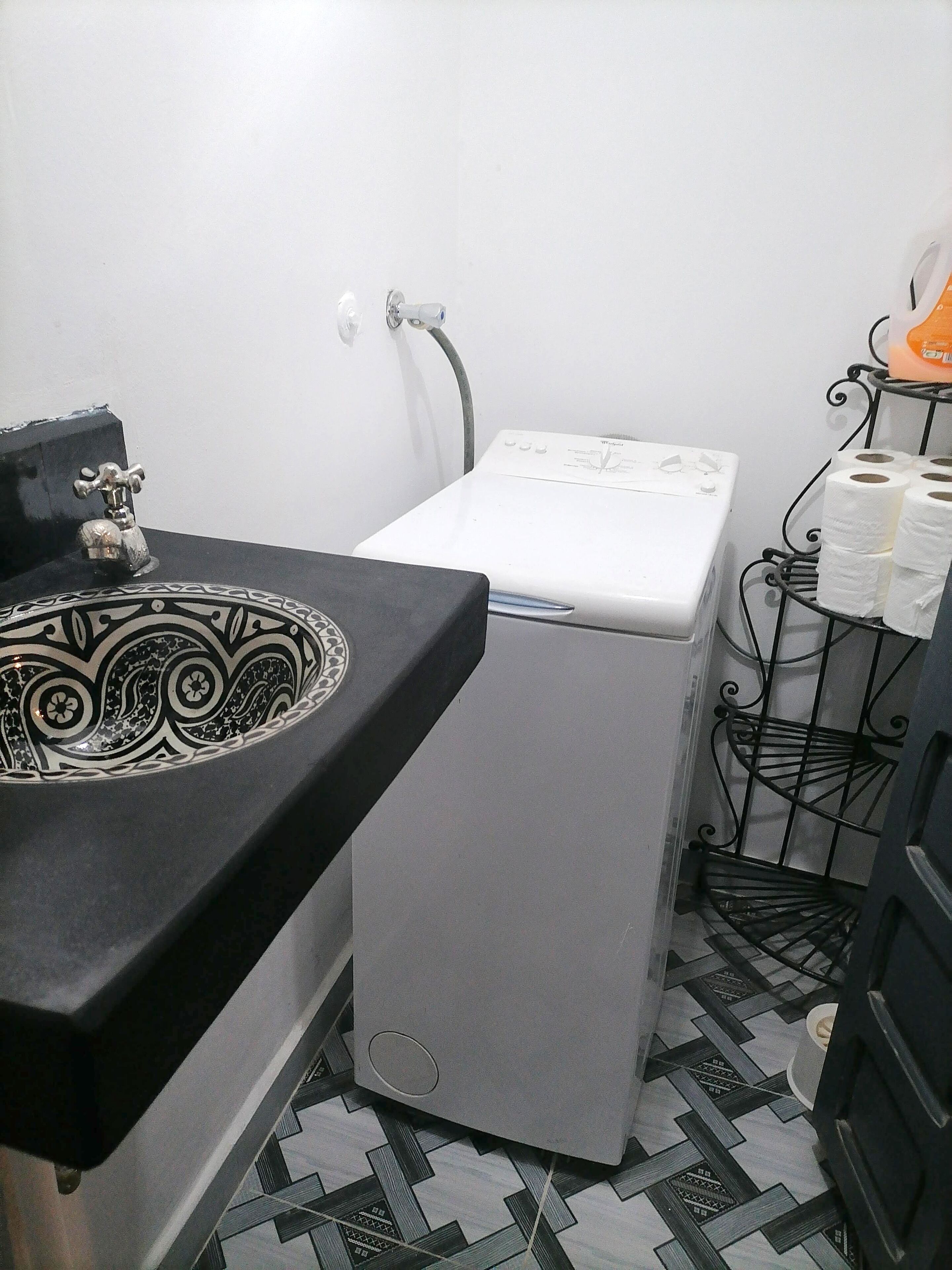 Un baño pequeño con un lavabo negro con un recipiente decorativo, una lavadora blanca y un estante metálico con papel higiénico.