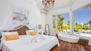 Un dormitorio amplio con una cama grande, una bañera y vistas a una piscina y paisaje tropical.
