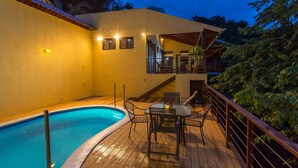 Una piscina con un deck de madera, una mesa y sillas, y un balcón con vistas a la piscina.