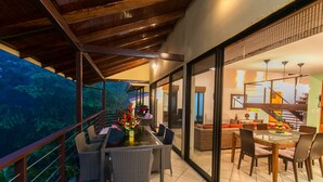 Comedor con mesa de vidrio y sillas de mimbre, con vistas a un jardín exuberante.