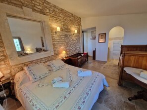Une chambre avec un mur en pierre, un grand lit avec une literie à motifs floraux, un banc en bois et une tête de lit en bois.