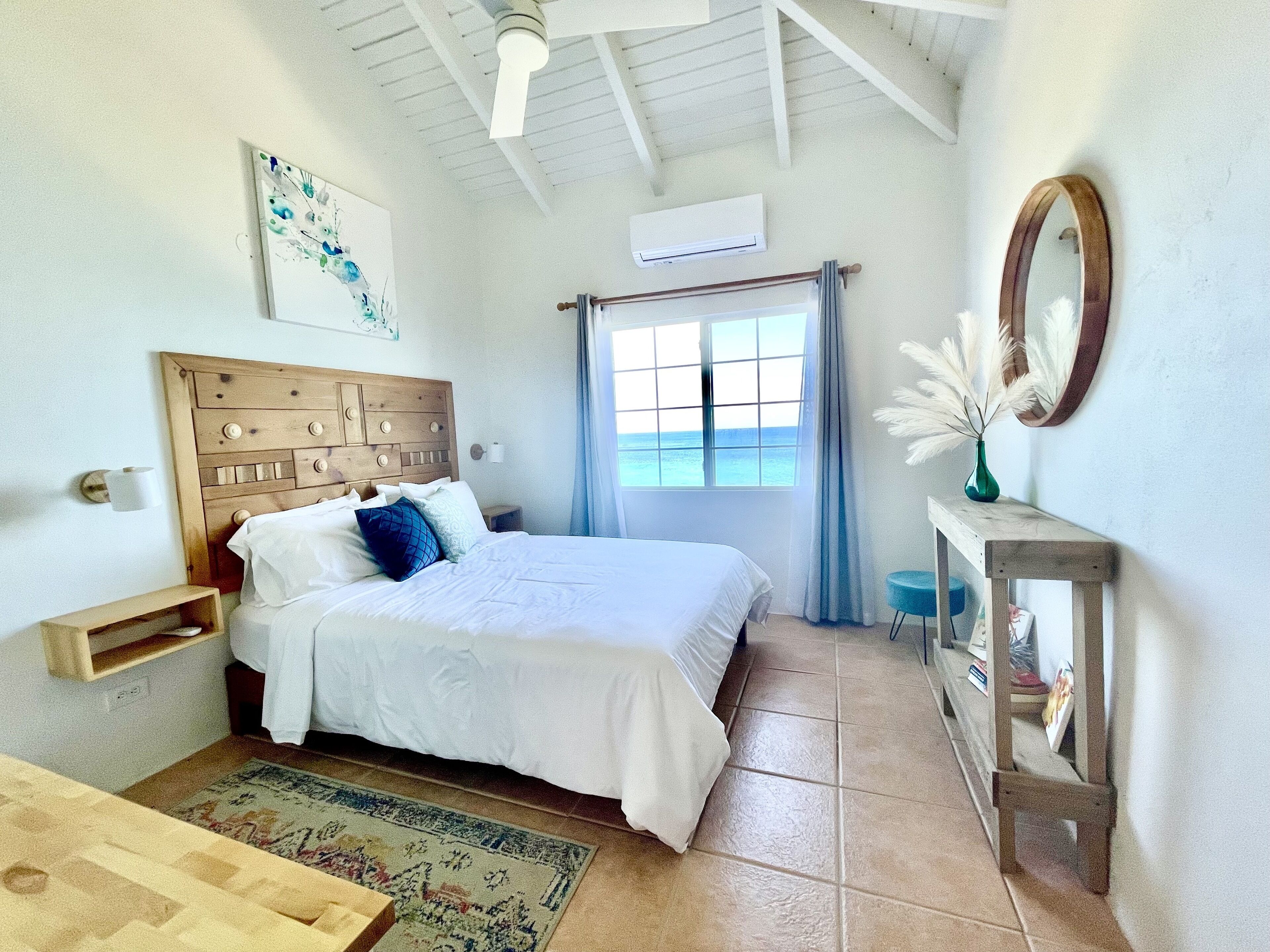 A bedroom with a wooden headboard, a bed with white bedding, a window with blue curtains, a mirror, and a small table with a vase.