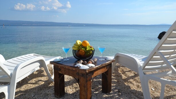 Een tafel aan het strand met een fruitschaal en twee martini glazen, omringd door witte lounge stoelen.