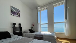 Een slaapkamer met twee bedden, een nachtkastje met een lamp, een schilderij en grote ramen met uitzicht op zee.
