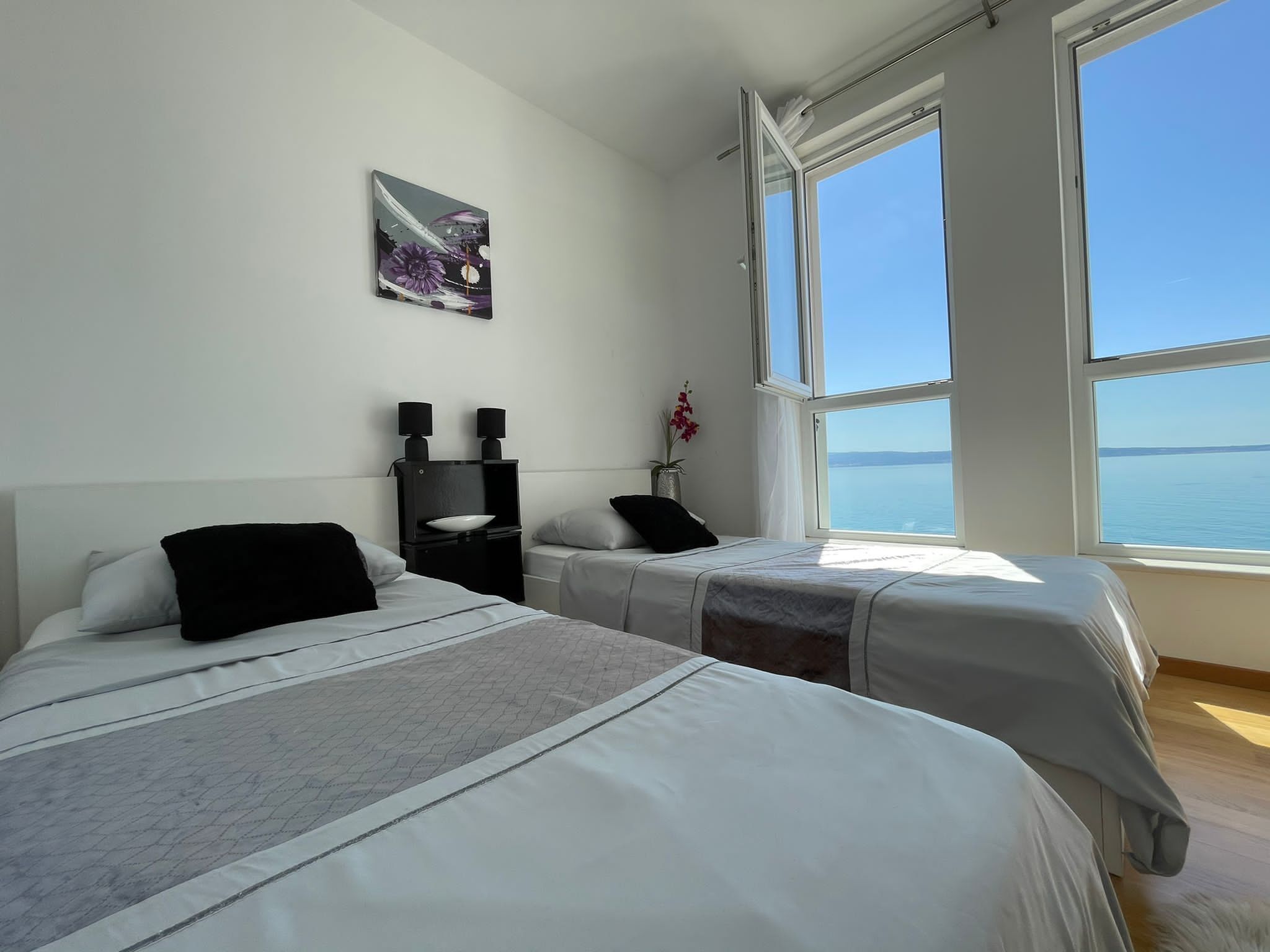 A hotel room with two beds, a nightstand with a lamp, a painting, and a large window offering a view of the sea.