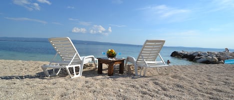 Una playa con dos sillas reclinables blancas, una mesa pequeña con un frutero y bebidas, y una costa rocosa al fondo.