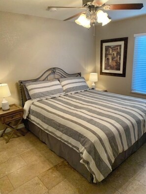 Une chambre à coucher comprenant un lit, une table de chevet, une lampe, un ventilateur de plafond et un tableau encadré accroché au mur.