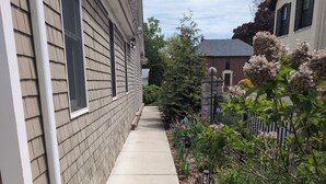 A narrow concrete pathway flanked by a building with beige siding and a fence with greenery.