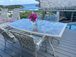 A glass-top table with patterned cushions, a vase of flowers, and a view of the ocean and houses in the background.