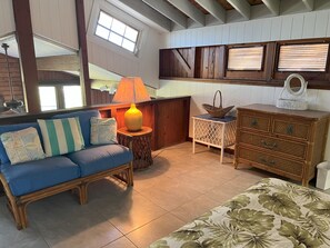 A cozy room with a blue sofa, a wicker side table with a lamp, a wicker basket, and a wooden dresser with drawers.
