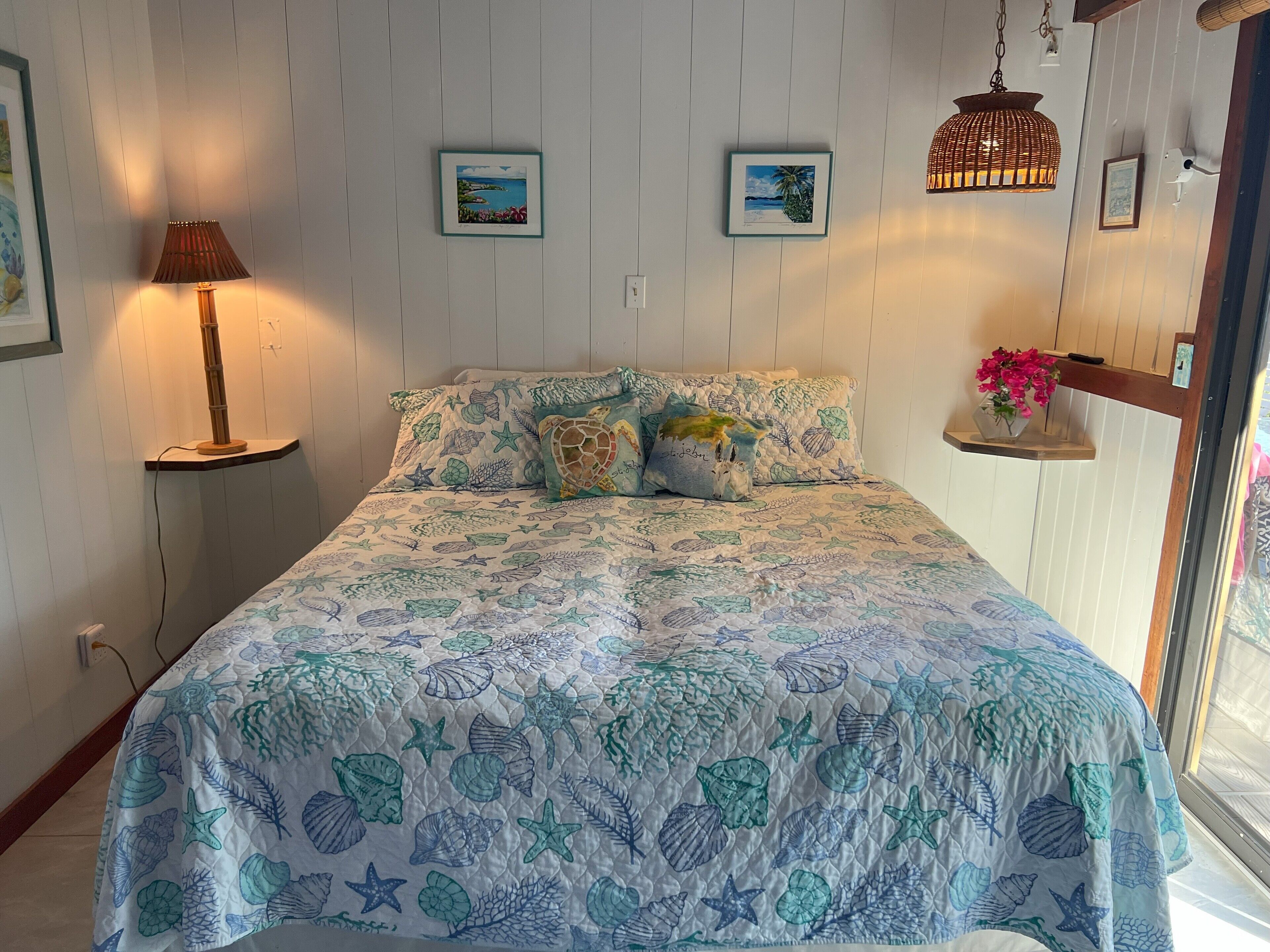 Un dormitorio con una cama adornada con un colcha de temática náutica, una lámpara de noche y un estante en la pared con un jarrón de flores.
