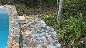 Un sendero de piedra que lleva a una piscina, rodeada de follaje y una terraza de madera.