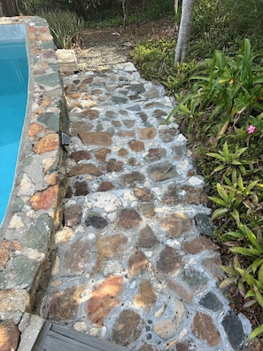 A stone pathway leading to a pool, surrounded by greenery and a wooden deck.