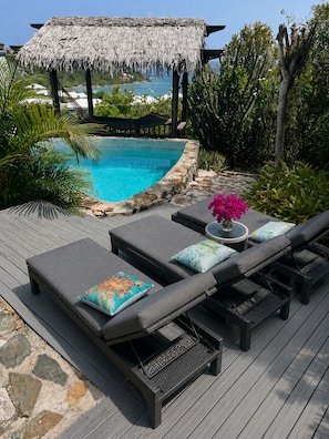 A poolside area with lounge chairs, a thatched roof, and a view of the ocean.