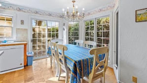 A dining area with a table set for four, surrounded by white chairs, a chandelier, and a view through large windows. - Vacation Home in Seabright (Santa Cruz)