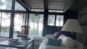 A room with a white sofa, a coffee table, and a lamp. There is a chair and a window with a view of trees. - Charlevoix Cottage: 150ft Lake Michigan Beach, Spectacular Sunsets & Dark Skies. (Charlevoix)