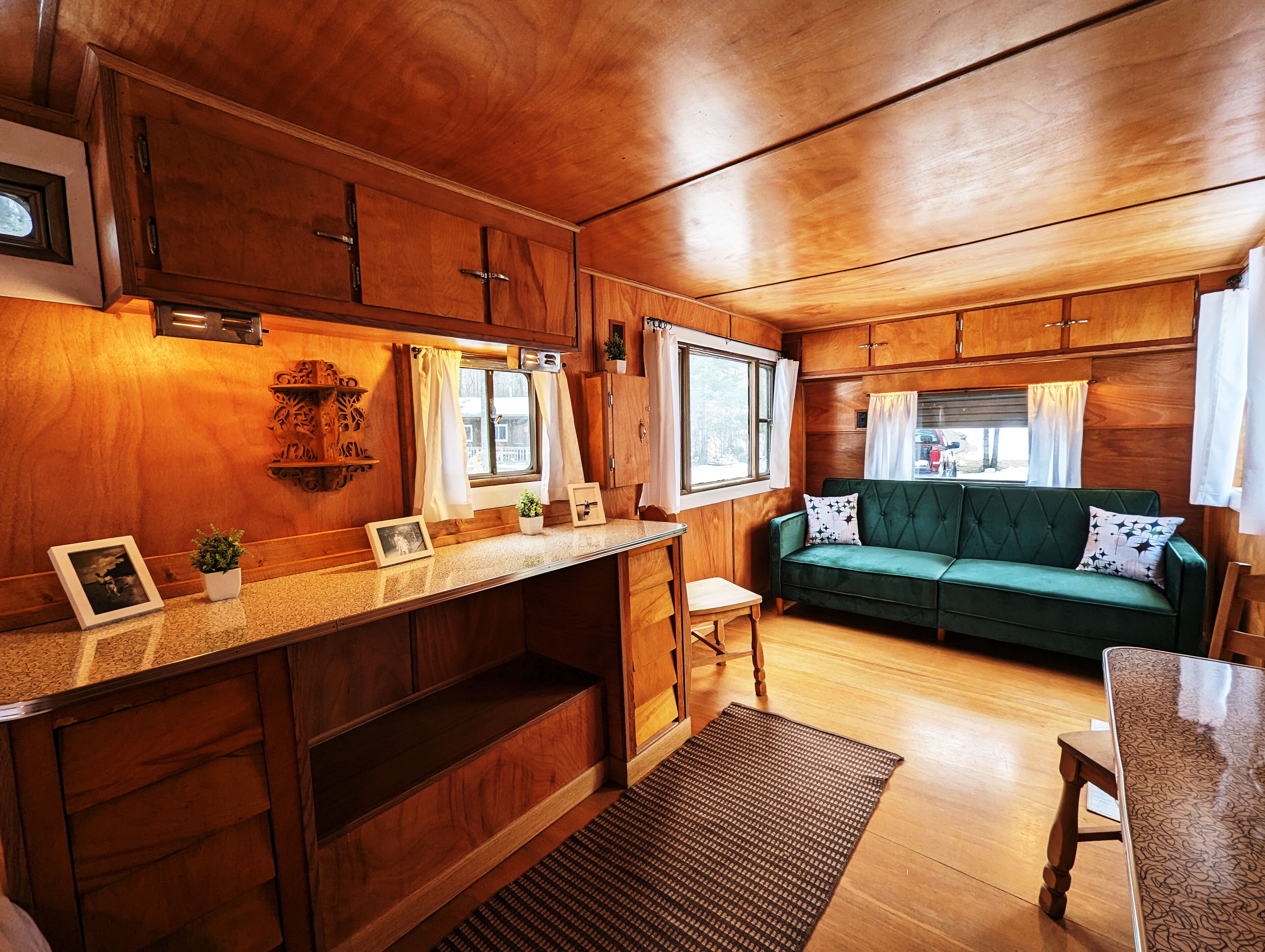 A cozy cabin interior with wooden walls, a green sofa, a small dining table, and a kitchen area with wooden cabinets and a window.