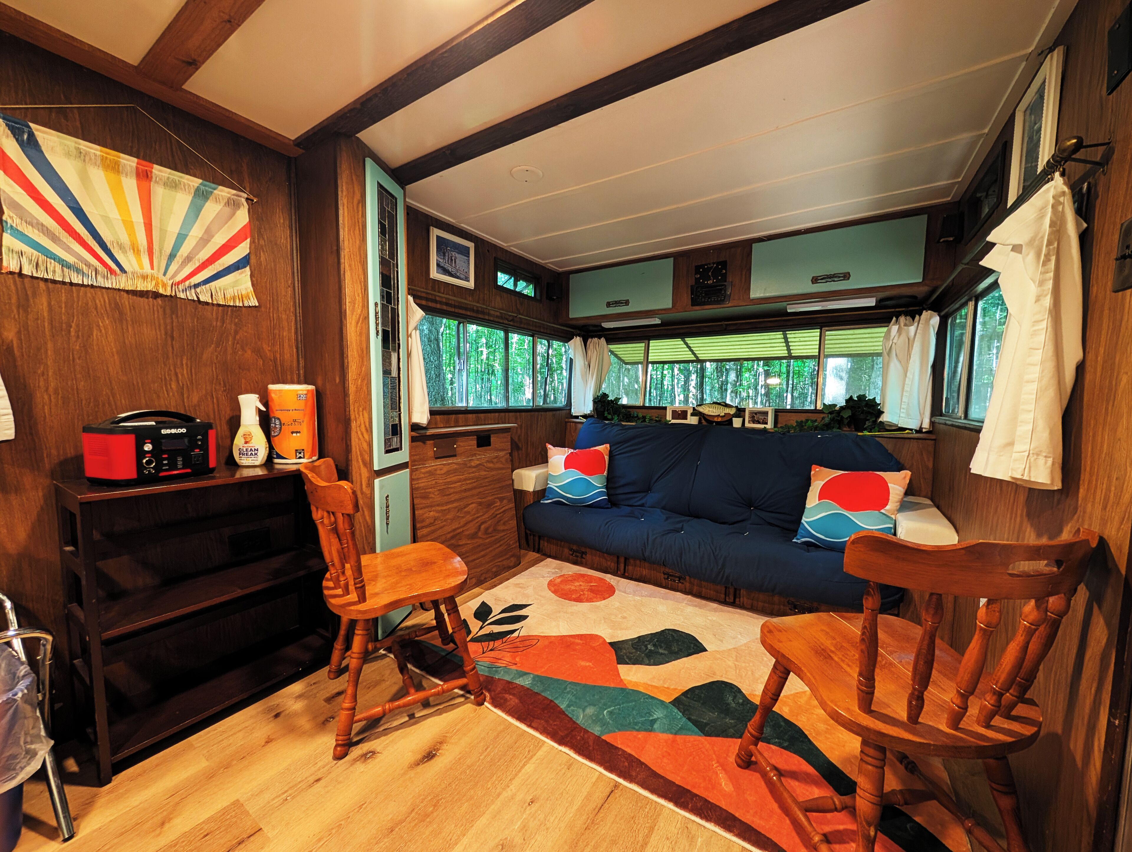 A cozy RV interior with a wooden floor, a blue sofa with colorful cushions, a wooden table, and a small kitchen area with a radio and cleaning supplies.