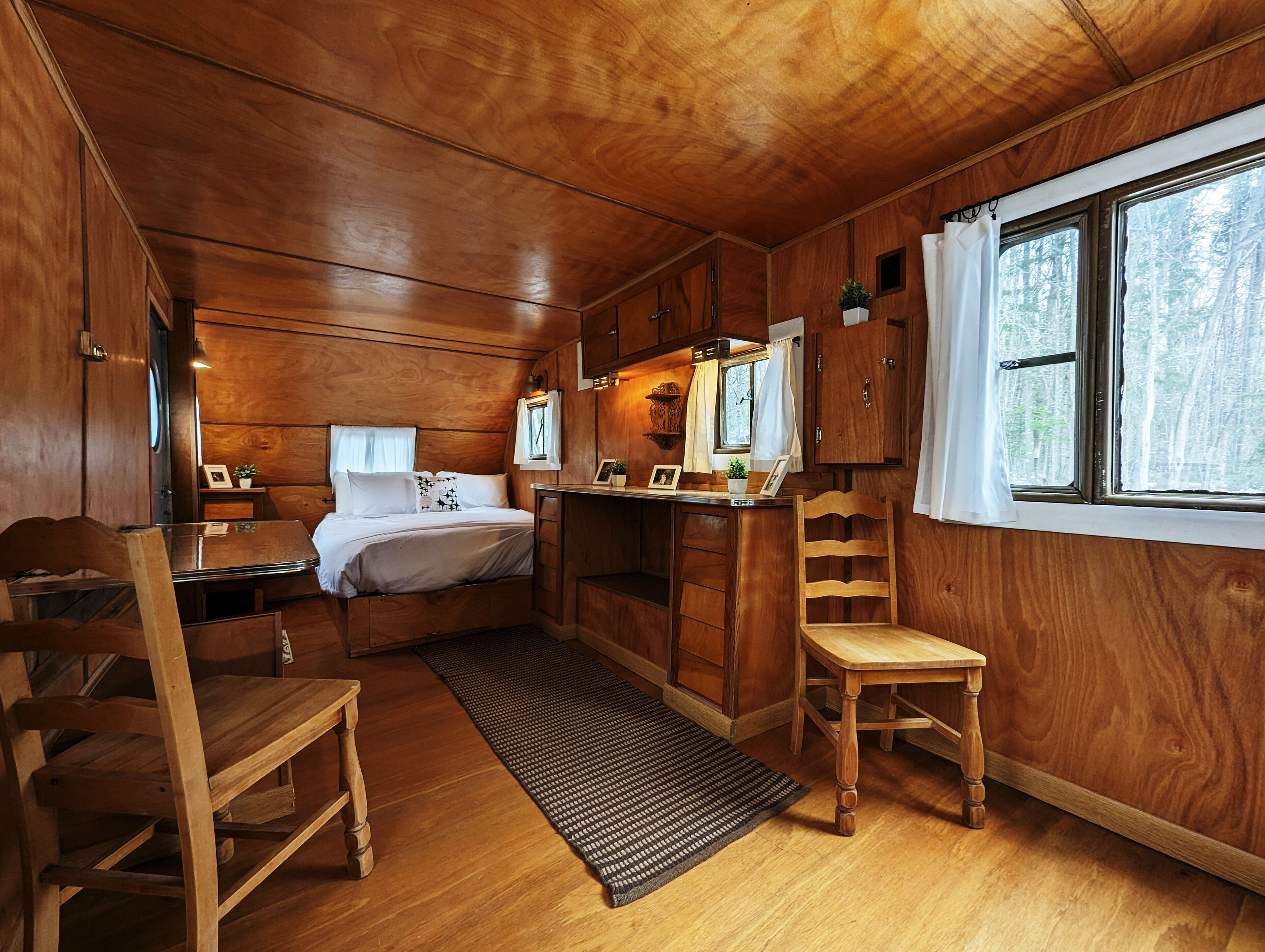 A cabin interior with a wooden interior, a bed, a desk, a chair, and a window with a view of trees.