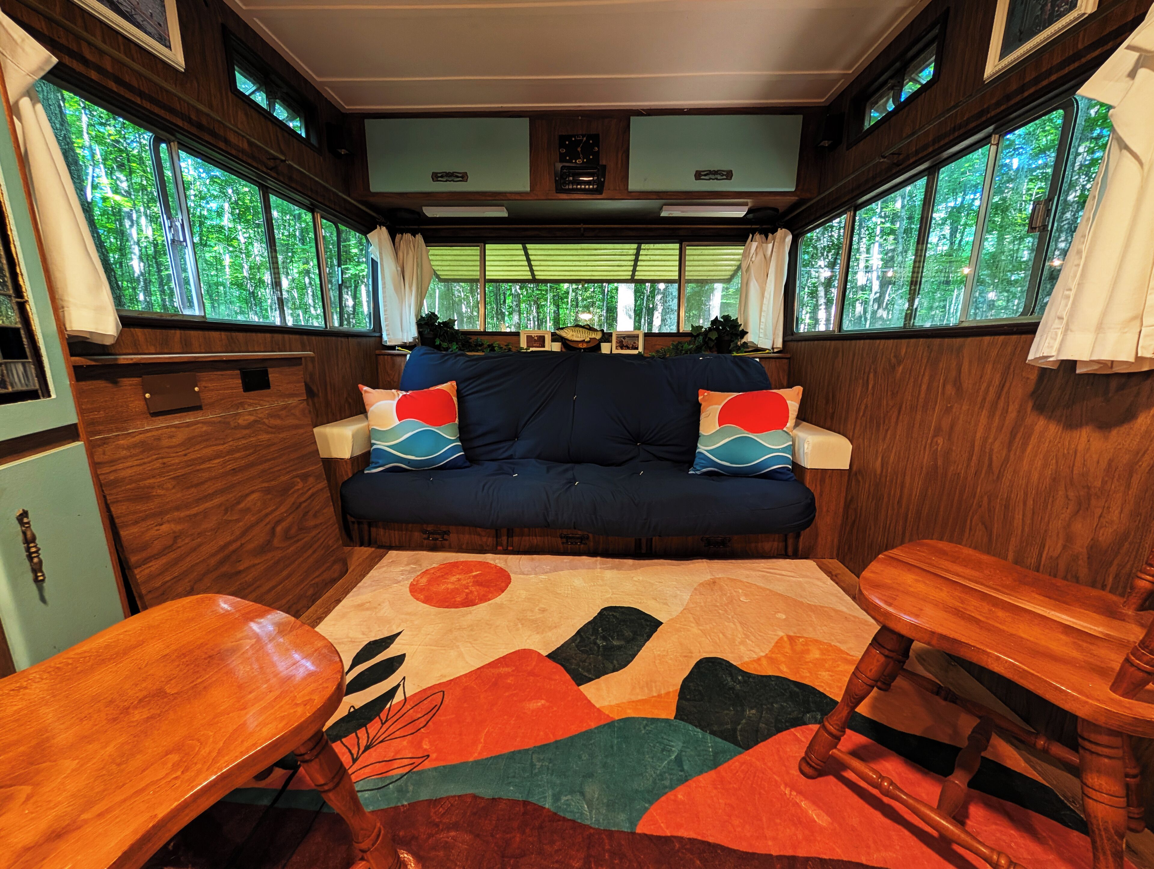 Interior acogedor de una autocaravana con un sofá, dos mesas de madera y un gran ventanal con vistas a un bosque.
