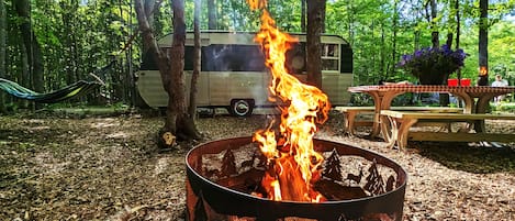 Una fogata en un bosque con una hamaca, una mesa con sillas y una tienda de campaña al fondo.