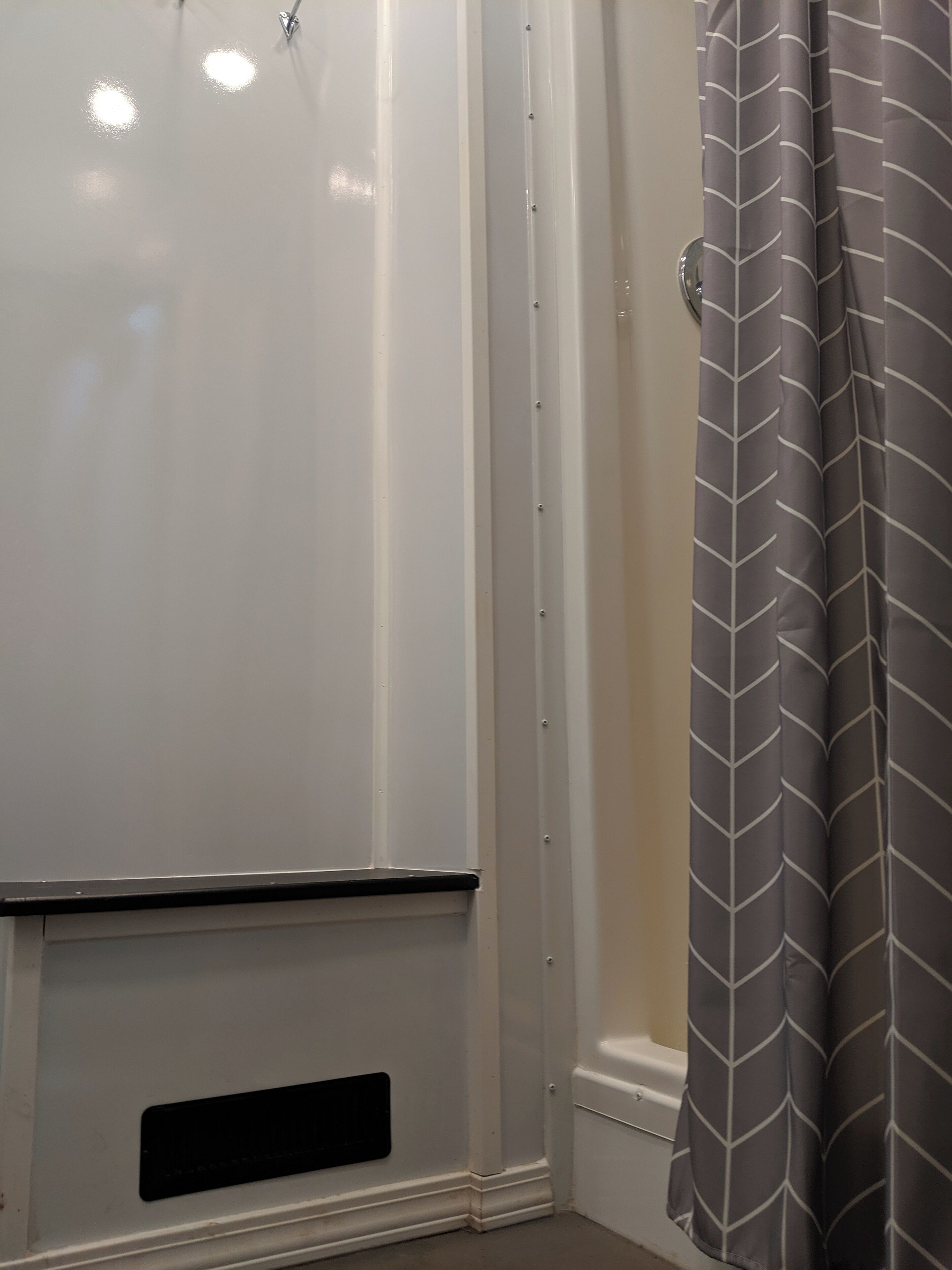 Une salle de bain avec une cabine de douche blanche, une grille d’aération noire et un rideau de douche gris à motifs.