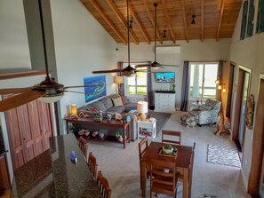 En rummelig opholdsstue med højt til loftet, træbjælker, ventilator, fjernsyn, sofa, sofabord og spisebord med stole.
