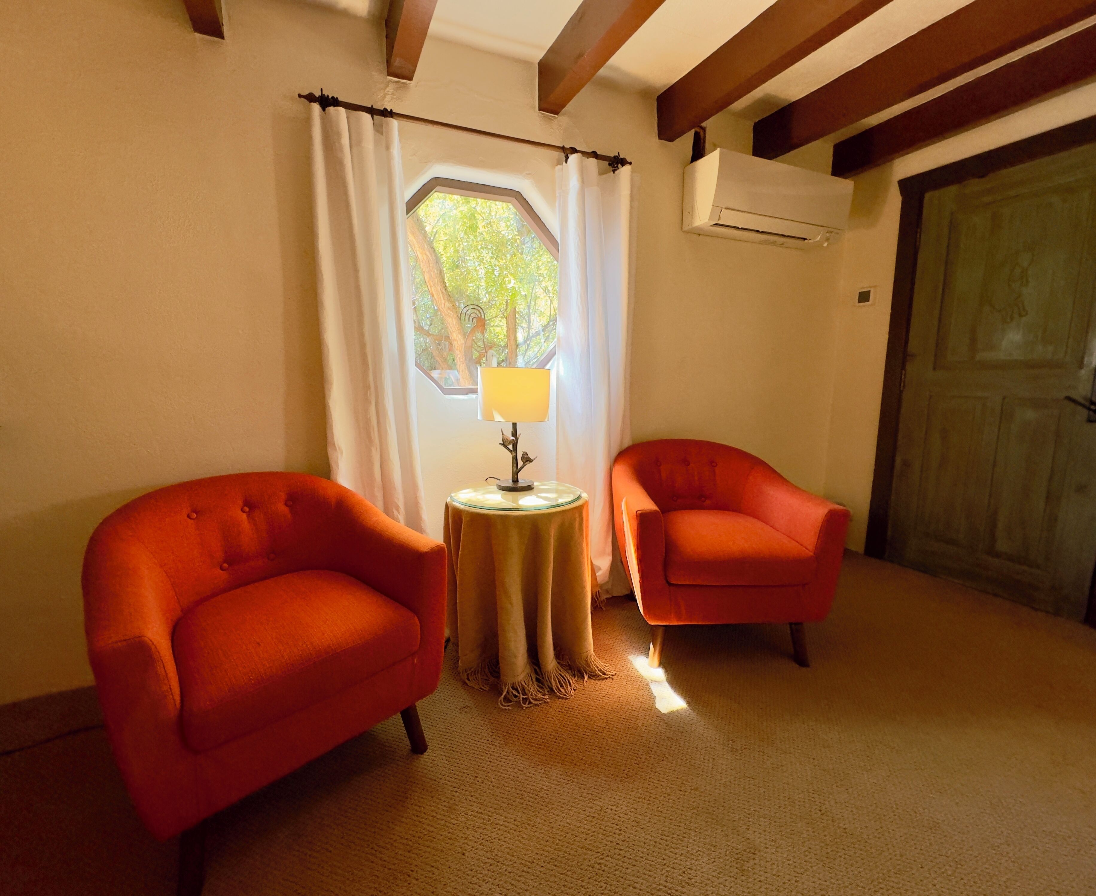 Une pièce avec deux fauteuils orange, une petite table ronde avec une lampe, une fenêtre avec des rideaux et un appareil de climatisation fixé au mur.