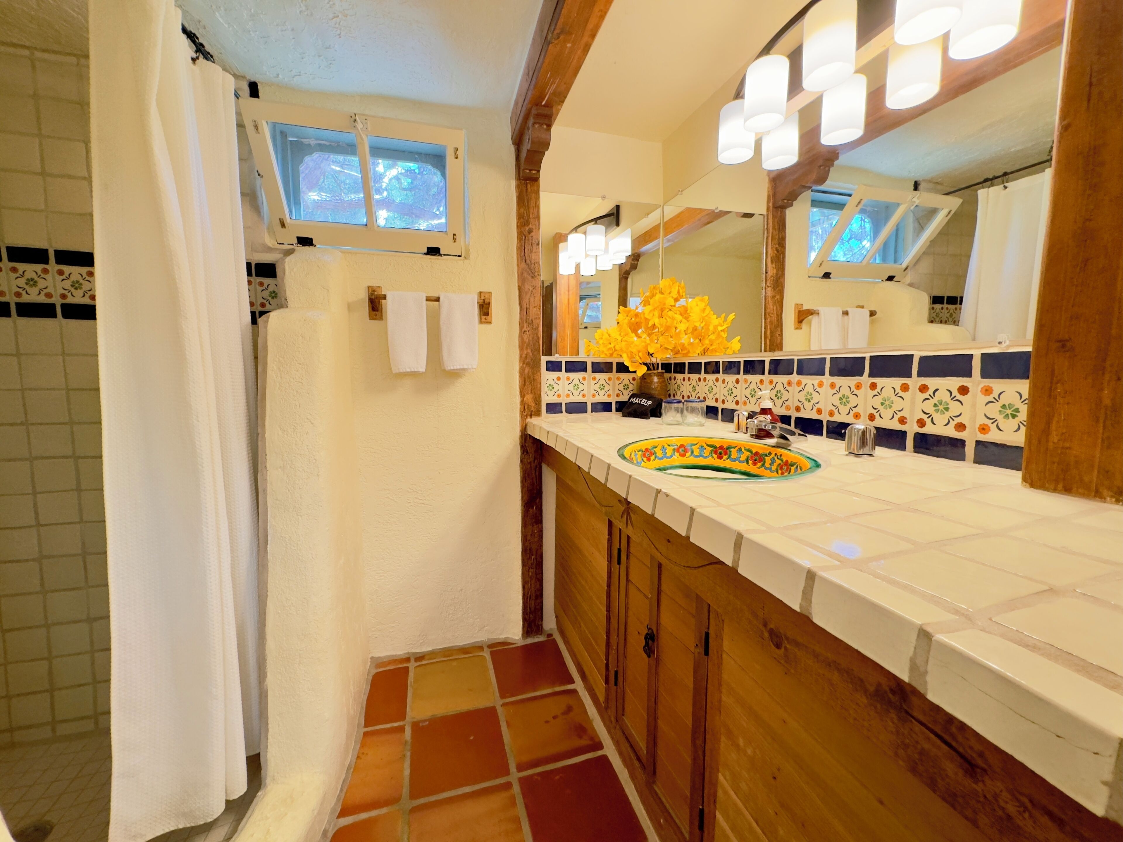 Un baño con piso de baldosas, tocador de madera, lavamanos con lavabo decorativo, espejo con iluminación y una ventana sobre el espejo.
