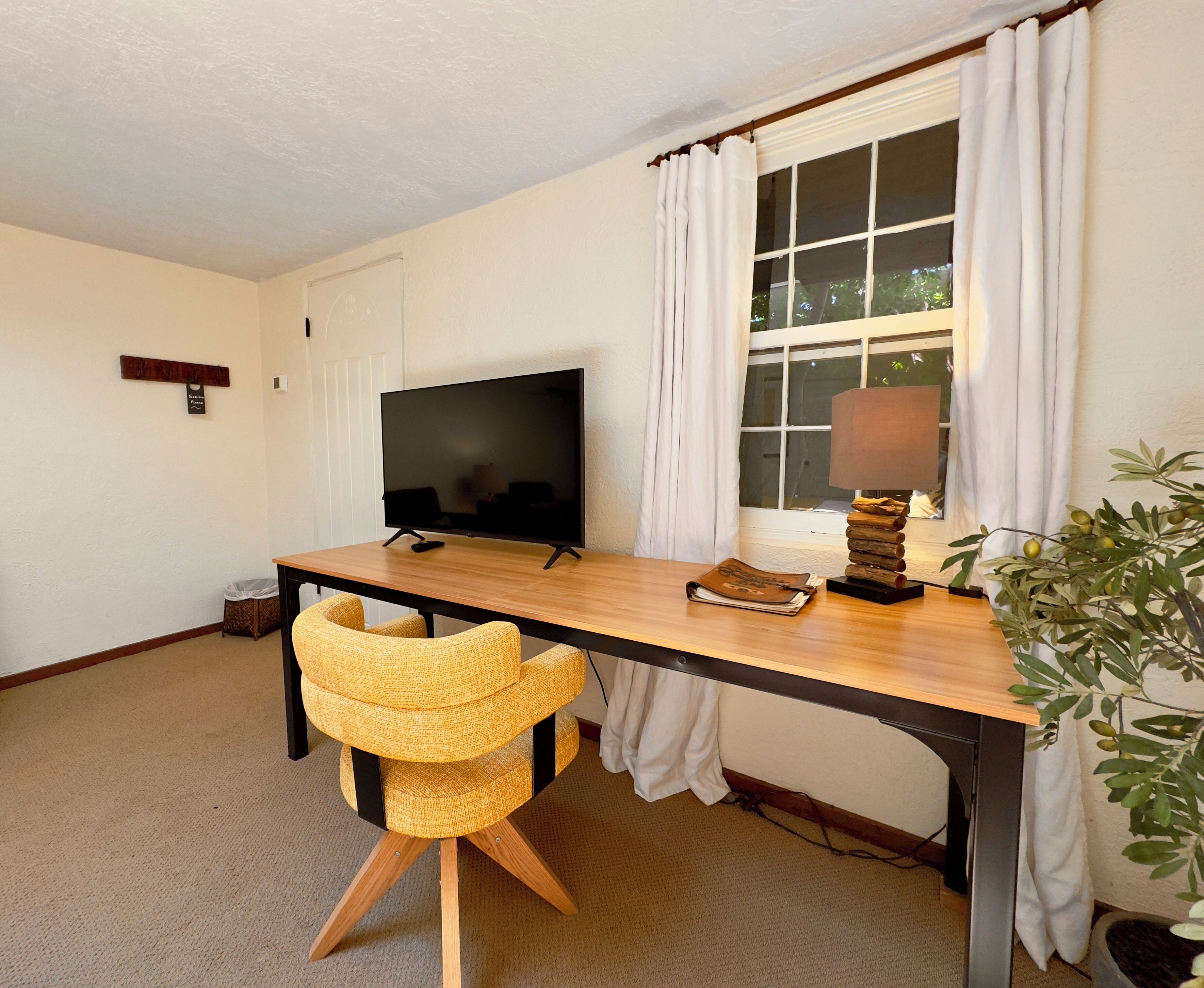 Ein Zimmer mit Holztisch, Fernseher, Lampe, Stuhl und einem Fenster mit Vorhängen.