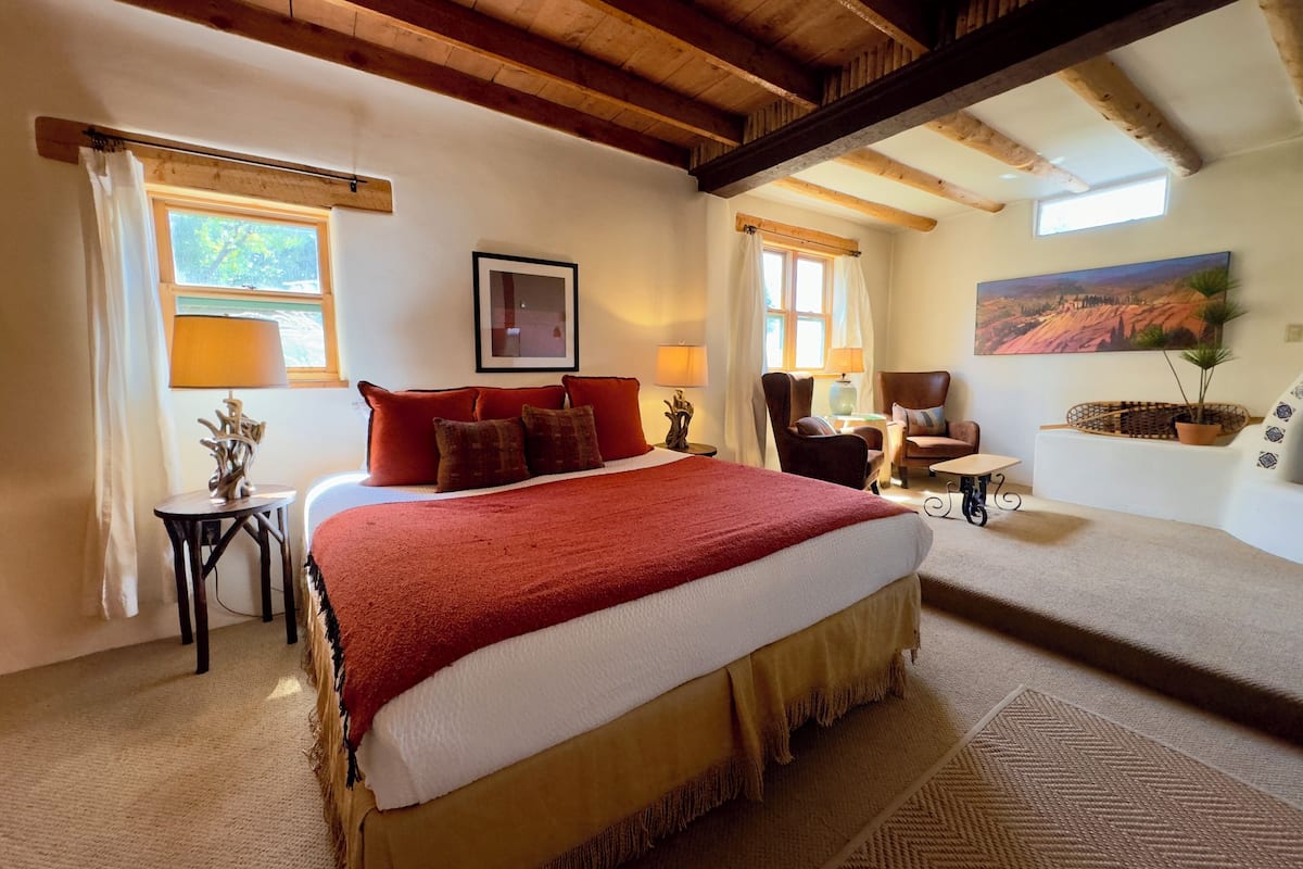Ruime slaapkamer met een groot bed, een bureau en een stoel. Er is een raam met gordijnen, een schilderij aan de muur en een plant op een plank.