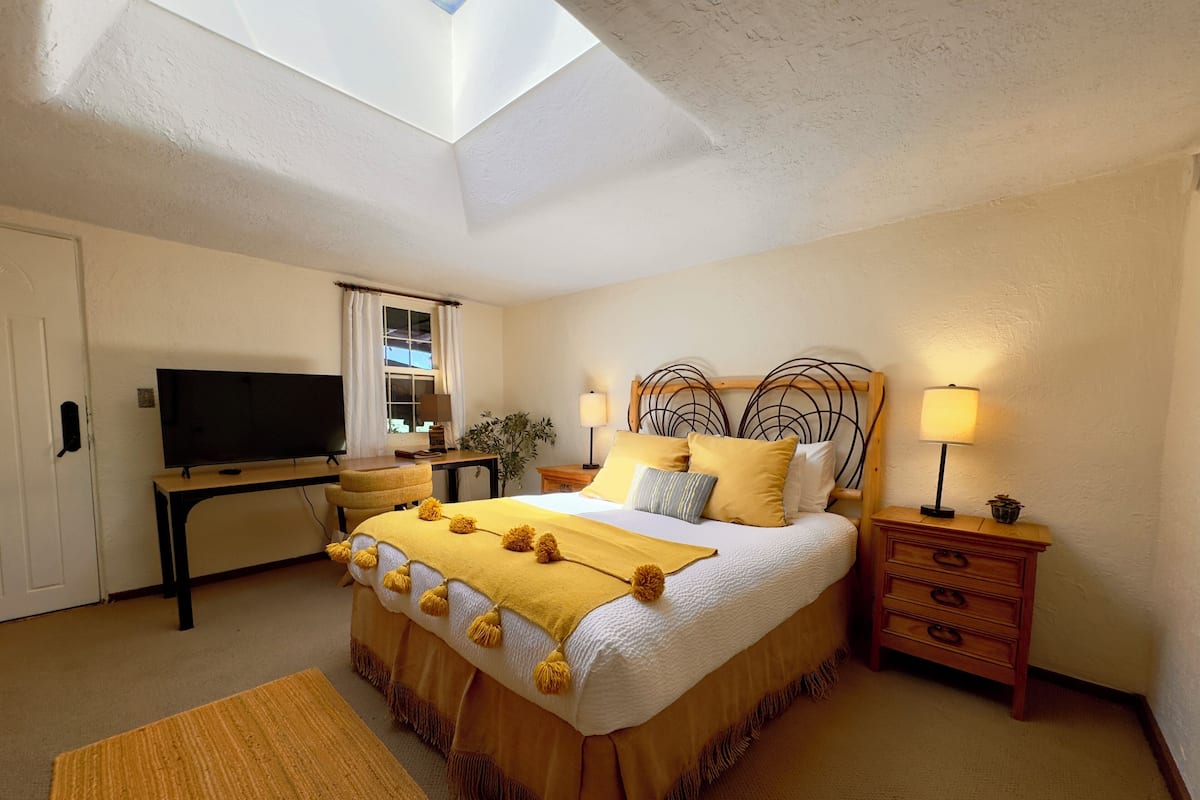 Een slaapkamer met een bed, nachtkastjes, een bureau, een televisie en een dakraam.