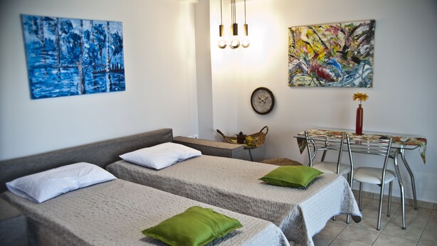 A room with two beds, a dining table with chairs, a clock, and two paintings on the wall. - Chalkida Apartments (Chalcis)