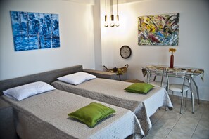 A room with two beds, a dining table with chairs, a clock, and two paintings on the wall. - Chalkida Apartments (Chalcis)