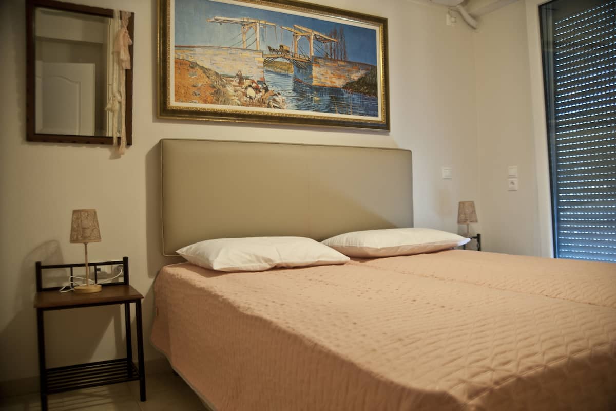 Un dormitorio con cama, mesita de noche, lámpara, un cuadro y ventana con persianas.