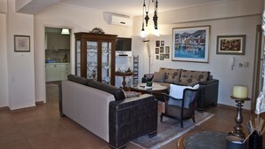 A living room with a sofa, armchairs, a coffee table, and a dining table. There is a painting on the wall, a chandelier, and a television in the background. - Chalkida Apartments (Chalcis)