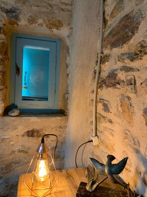 A rustic stone wall with a wooden table, a hanging light fixture, and a small window with a blue frame. - Nomitsi Traditional House (West Mani)