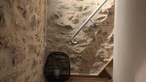 A staircase with a stone wall on one side and a wooden floor. A metal handrail is present, and a black woven basket is placed on the floor near the bottom of the stairs. - Nomitsi Traditional House (West Mani)