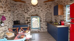 A rustic stone-walled room with a dining table set for two, a kitchen area with a red refrigerator, and a window with a view of a tiled roof. - Nomitsi Traditional House (West Mani)