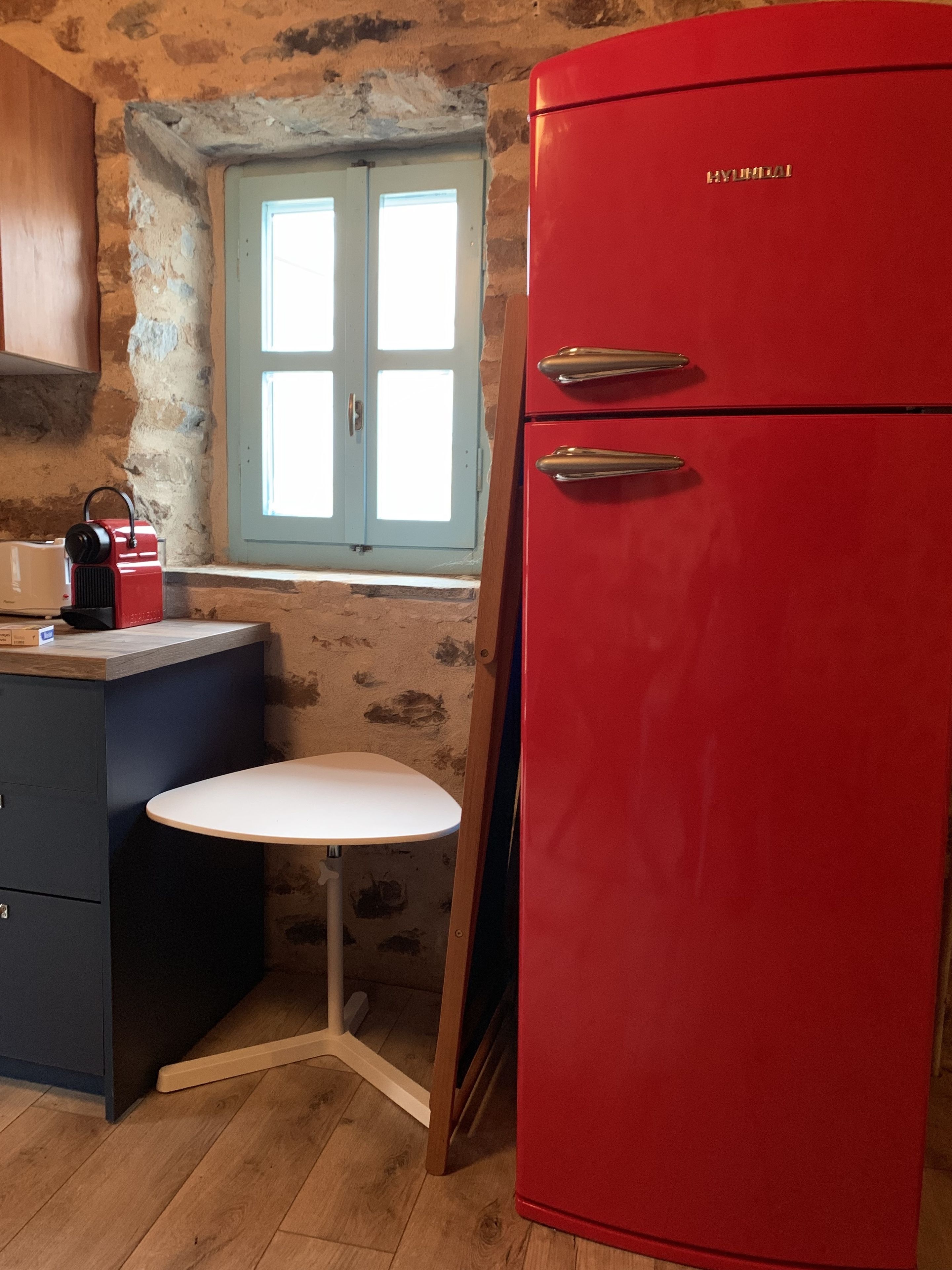 A compact kitchen with a red refrigerator, a small white table, and a window with blue shutters.