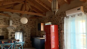 A rustic kitchen with stone walls, a wooden ceiling, a red refrigerator, and a dining area with blue chairs. - Nomitsi Traditional House (West Mani)