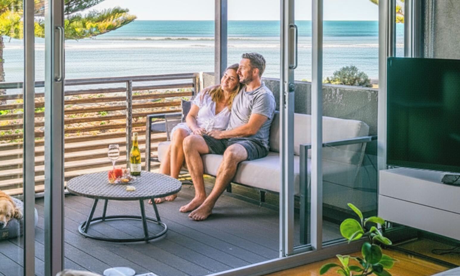 A couple seated on a balcony with a view of the sea, a table with a bottle, and a TV on a stand.