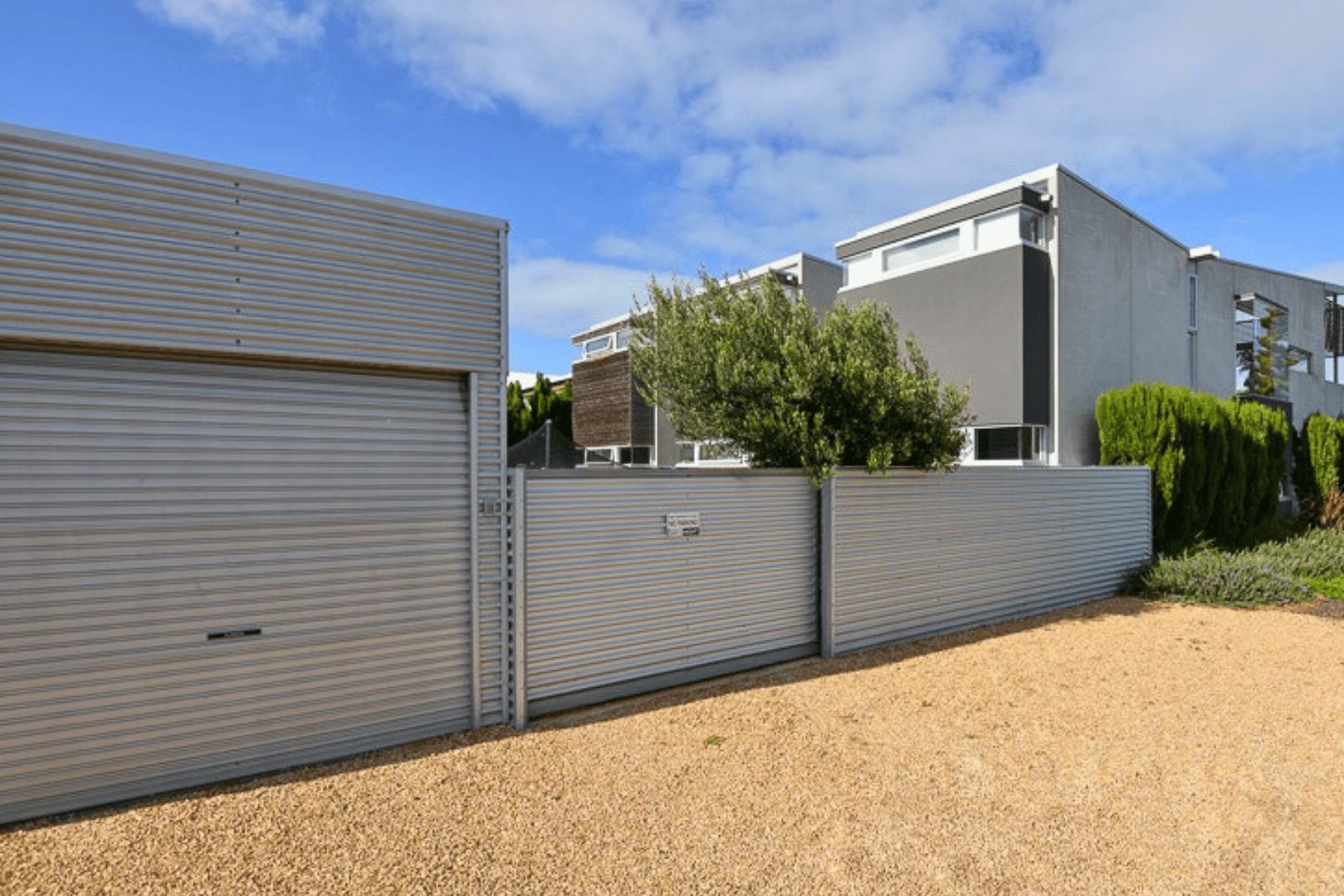 A modern building with a metal fence, a gravel driveway, and a clear sky.
