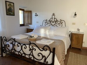 Una cama bien hecha con cabecera metálica, una mesita de noche con lámpara y un pequeño armario de madera.
