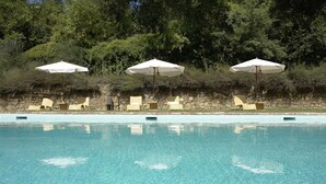 Una piscina con agua azul cristalina, rodeada de muros de piedra y protegida por sombrillas blancas.