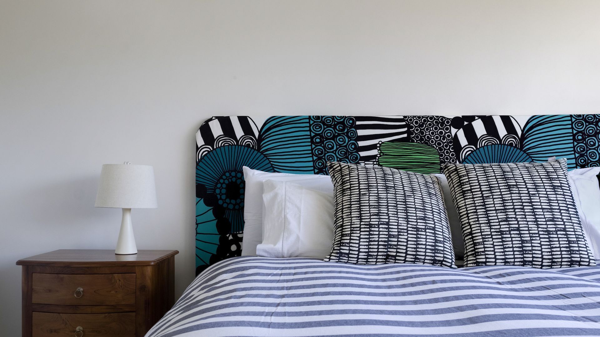 A bed with striped bedding, a patterned headboard, and a bedside table with a lamp.