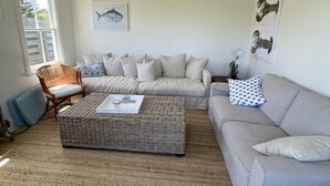 Un salon comprenant un grand canapé d’angle, un pouf en osier, une chaise en bois et un lampadaire. Les murs sont ornés d’œuvres d’art et le plafond est équipé d’un ventilateur.